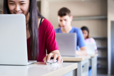 In a high school classroom a group of cheerful students study using a laptop computer. A happy young Asian girl looks at the screen. Concept of technology and education. Focus on the mouse.