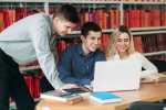 University students sitting together at table with books and laptop. Happy young people doing group study in library